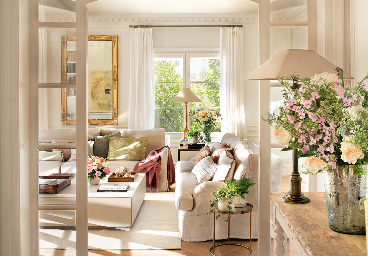 Recibidor cl&aacute;sico con sal&oacute;n al fondo, puerta con arco de cristal, sof&aacute;s blancos y flores